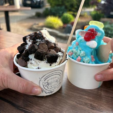 Two hands holding paper cups of ice cream outdoors—one vanilla topped with brownie bites and cookies, and the other bright blue with colorful candy toppings