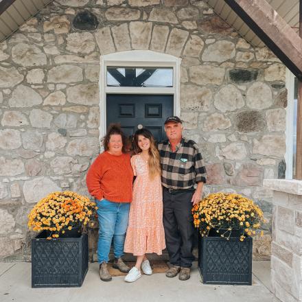 three people stand on a farmhouse porch in between potted mums