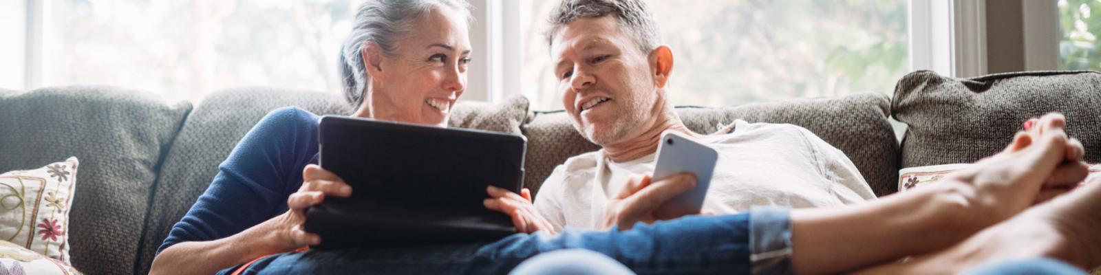 Woman and man seated closely on couch looking at at tablet and cell phone