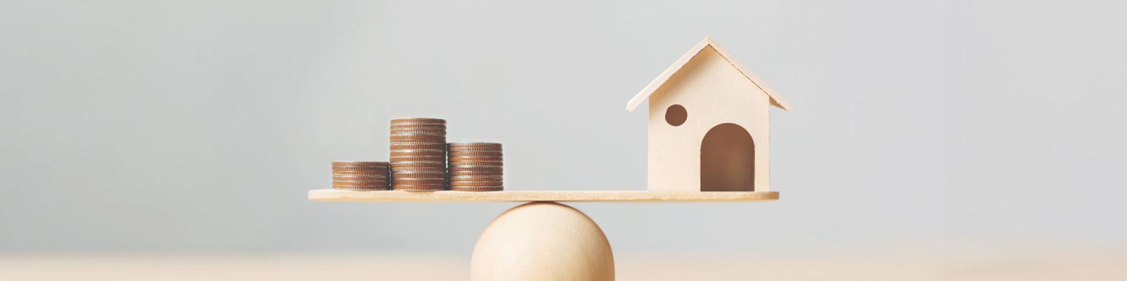 A miniature wooden house balancing on a plank next to stacks of coins, sitting on a wooden ball