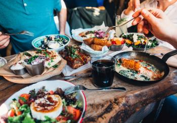 a rustic wooden table filled with various Mediterranean-style dishes, including salads with goat cheese, roasted vegetables, and grilled meats, being shared by a group of people