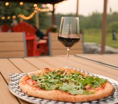 woodfired pizza topped with arugula and a glass of red wine on a light-colored wood-slat table in an outdoor setting.