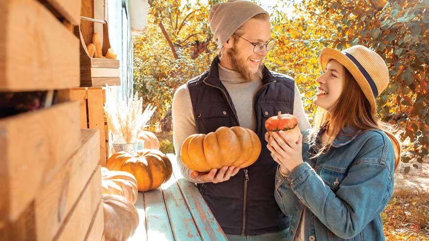 A man in a toque and woman in straw hat smiling at each other holding miniature pumpkins at an outdoor market and pumpkin patch