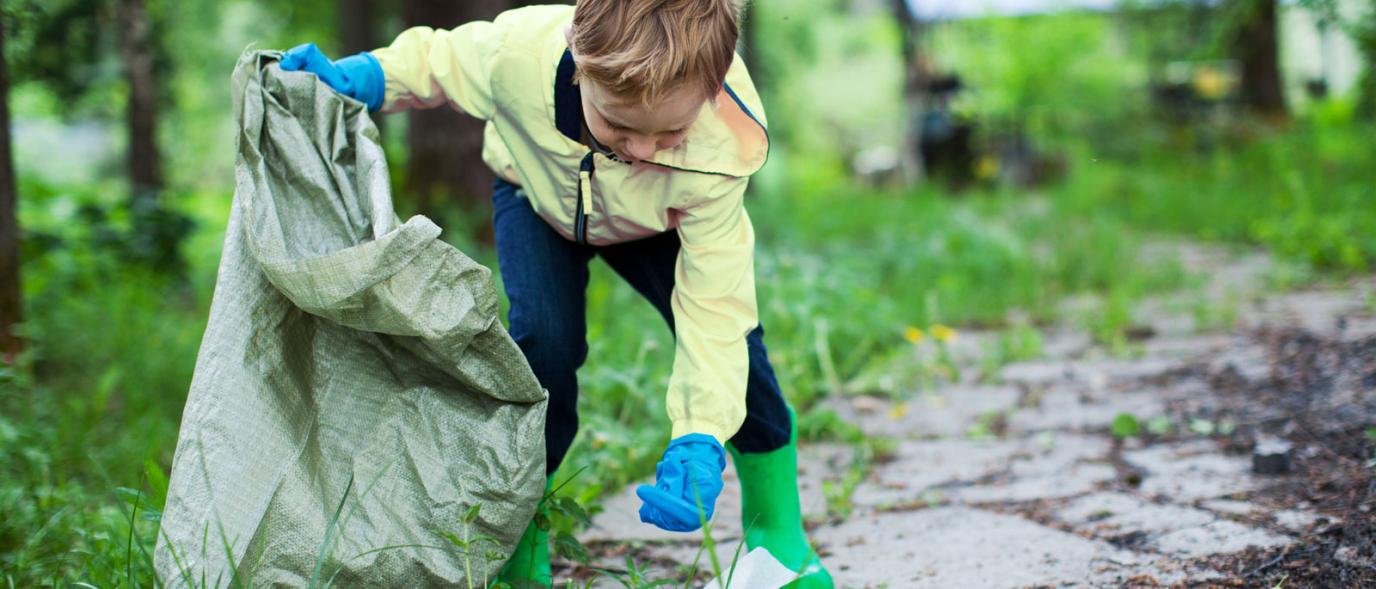 Boy picking up garbage in a park setting wearing gloves, boots and a raincoat