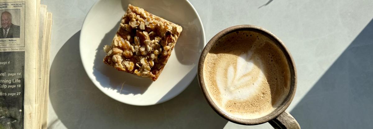 A latte and a square pastry on a table in the sunlight.