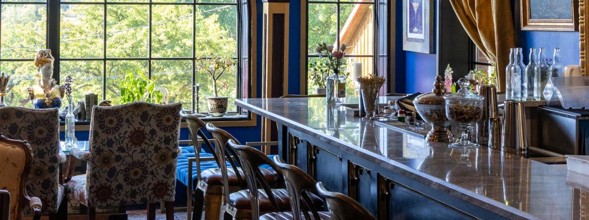 An elegant interior bar with blue walls, large windows, bar seating, and dining chairs.