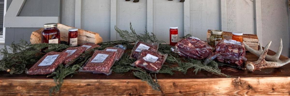 Packaged meats and artisan goods displayed on a wooden table with evergreen branches and deer antlers.
