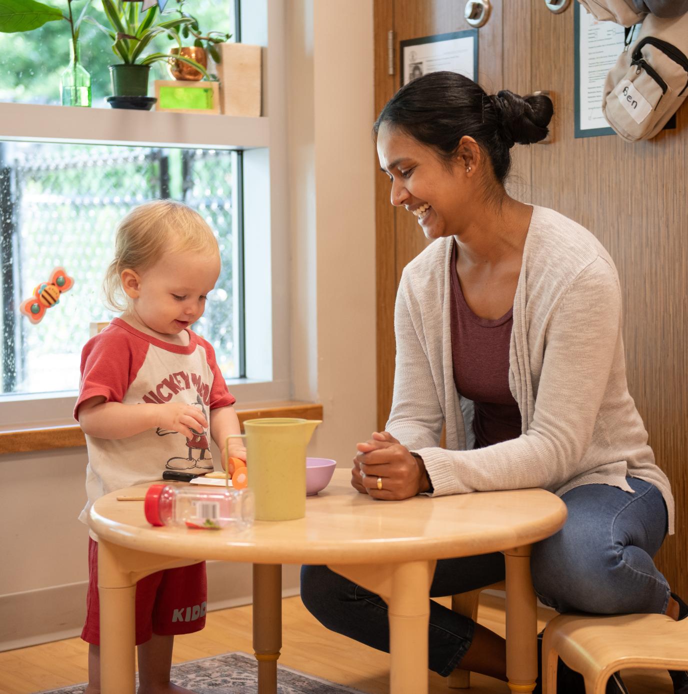 A child care worker sits at a table smiling at a small child. 